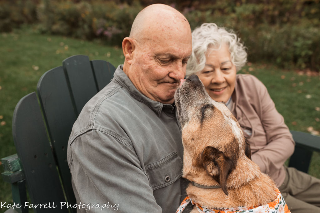 Red Heeler licks Grandpa's nose while he sits next to Grandma in Adirondack chairs.