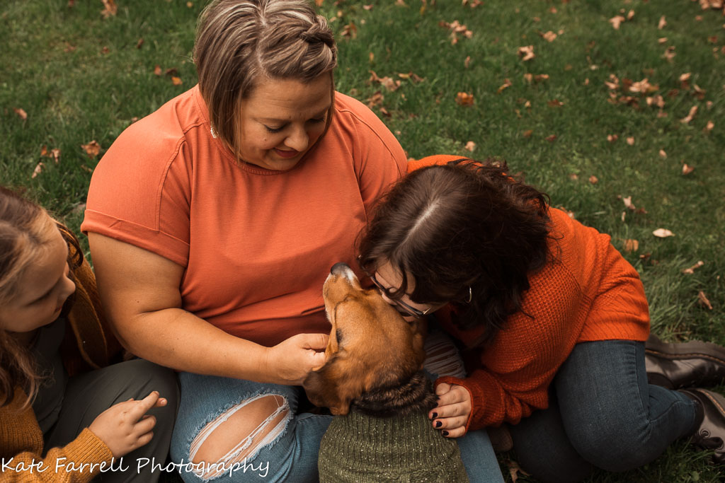 Mom and two daughters snuggle with their dog in the grass.