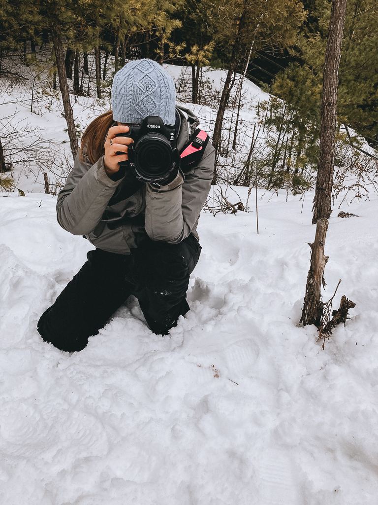 A woman in a blue hat kneels in the snow to take a photo.