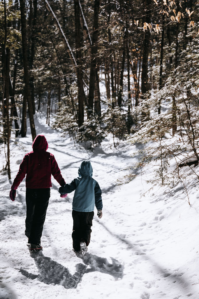 Two girls photographed from behind walking on a snowy trail in the woods.