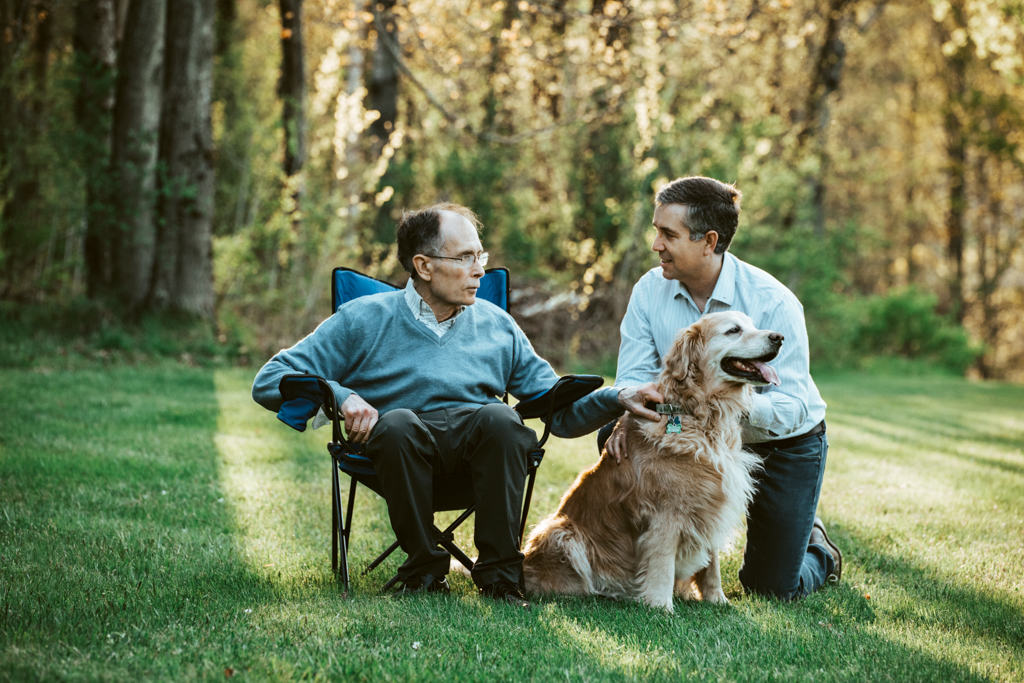 A man and his elderly father visit on a sunny spring afternoon while petting a golden retriever.