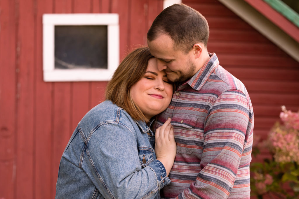 A young couple hugs in front of a red barn.
