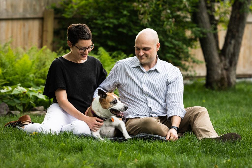 A man and woman snuggle with their dog in the grass in the spring in their backyard.