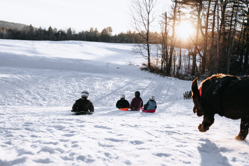 Four children zoom down a sledding hill with a young dog chasing them.