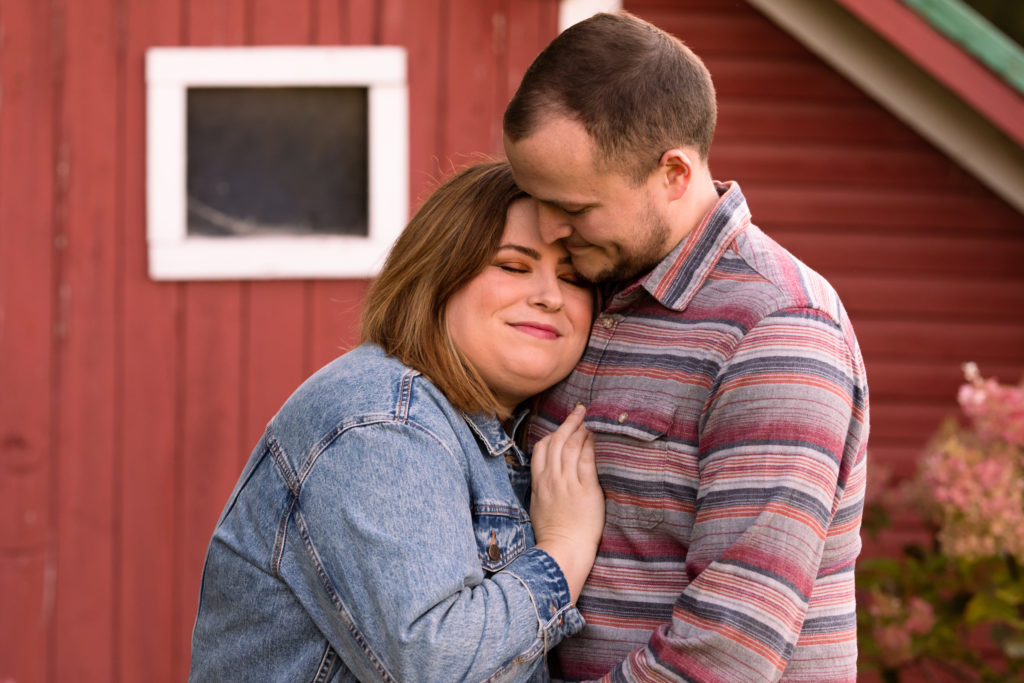 A couple in their 20s hugging in front of a red barn in northern Vermont. 