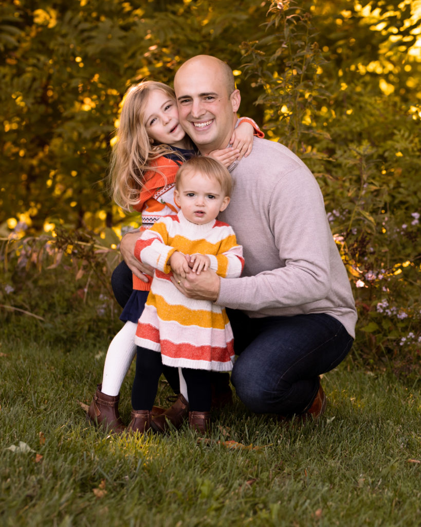 Dad snuggles his two little girls with fall colors behind them.