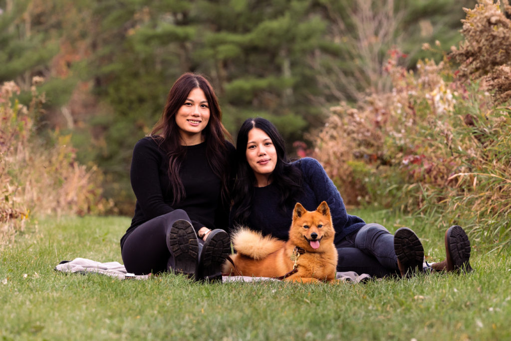 Two women sit with their dog on the grass with fall colors in the background.