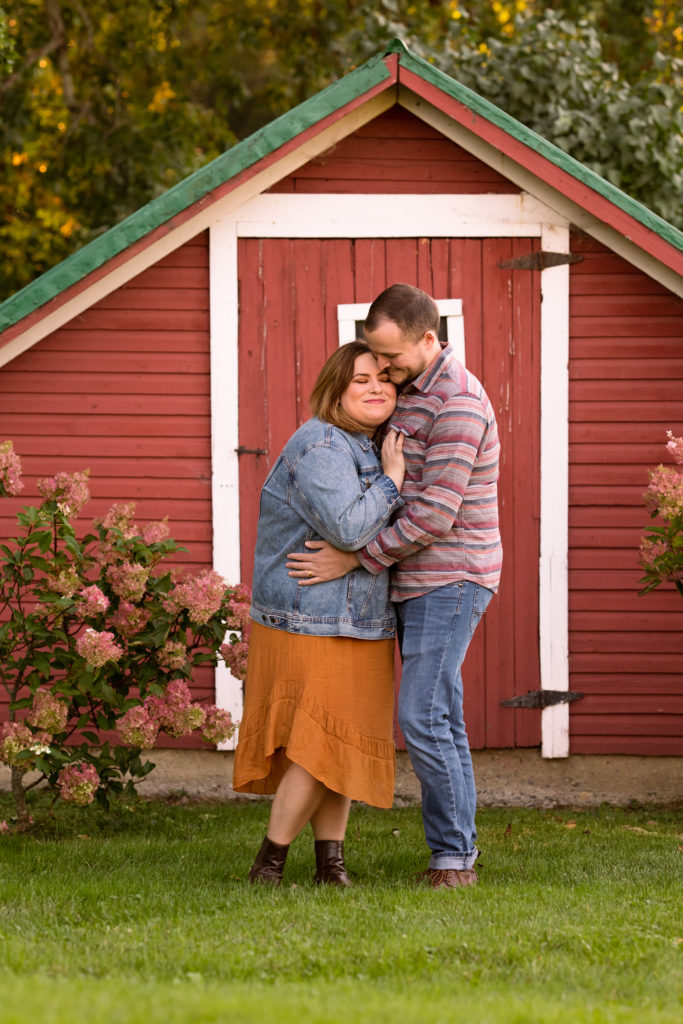 A young couple hugging in front of an old red shed - Vermont Outdoor Family Photography