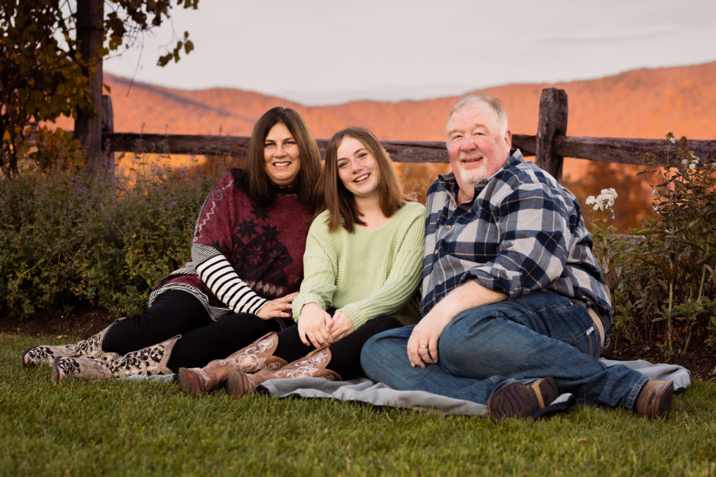 Parents and their teenage daughter relax on a blanket in front of a fence and fall foliage - Vermont Family Photography 