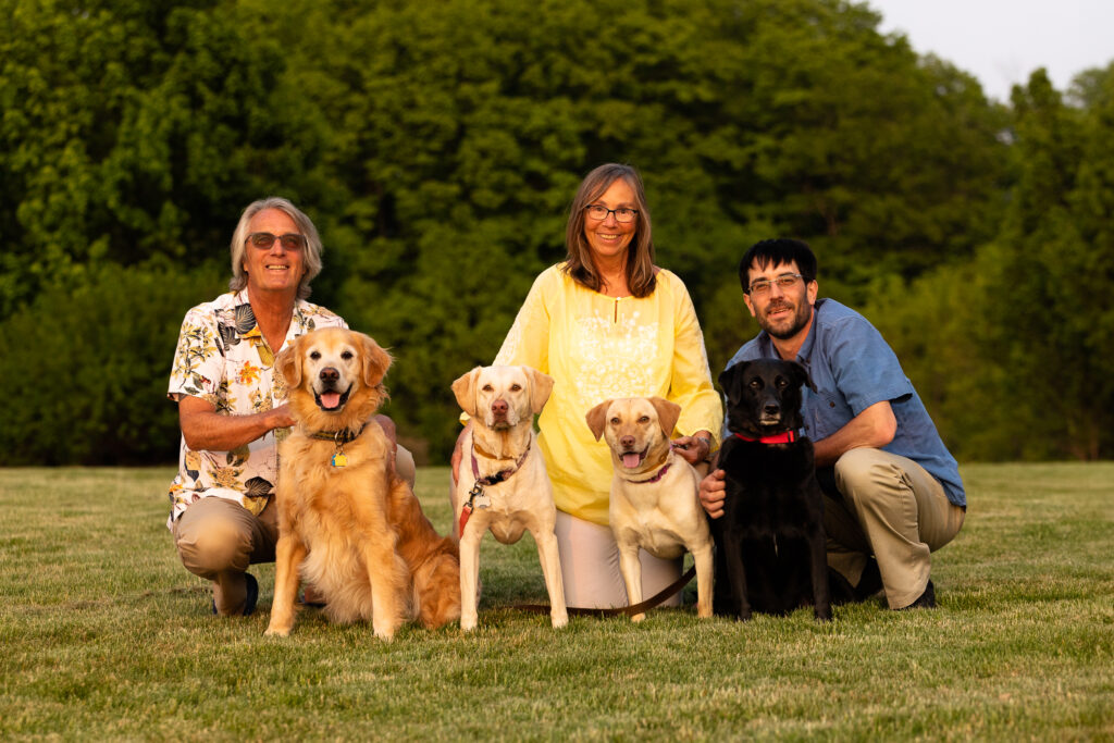 Three adults and four dogs in the grass with a green trees behind them. Vermont dog photography.