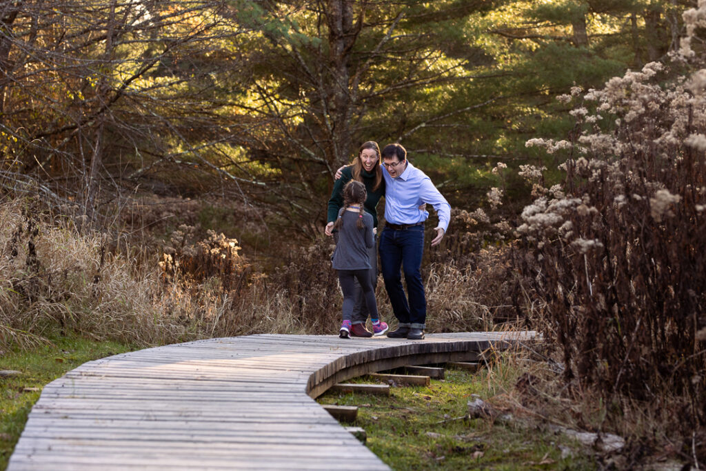 A seven year old runs up to Mom and Dad on a wooden bridge in the woods to surprise them with a hug.